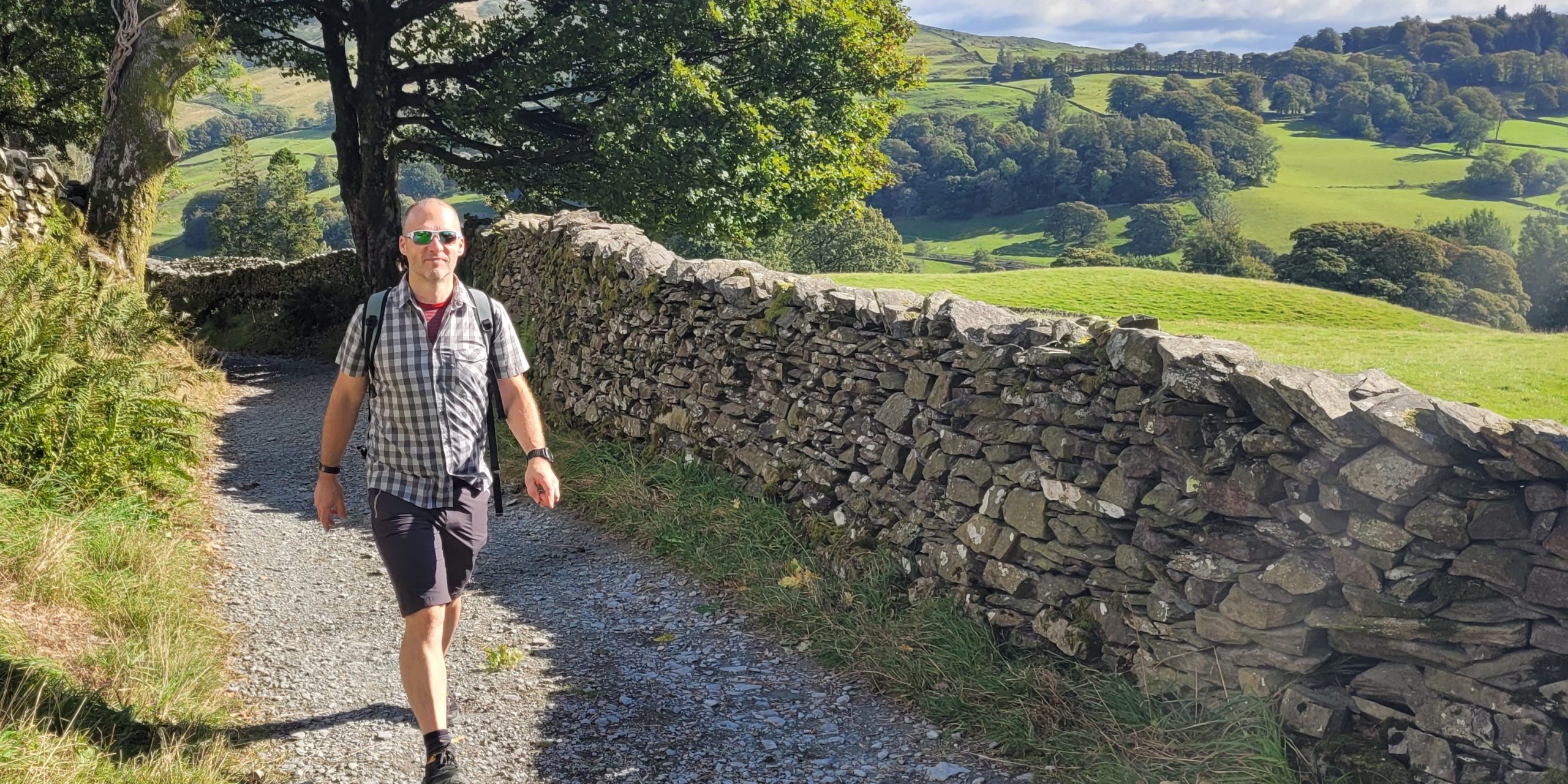 Man walking on a countryside path alongside a dry stone wall, with rolling hills in the background.